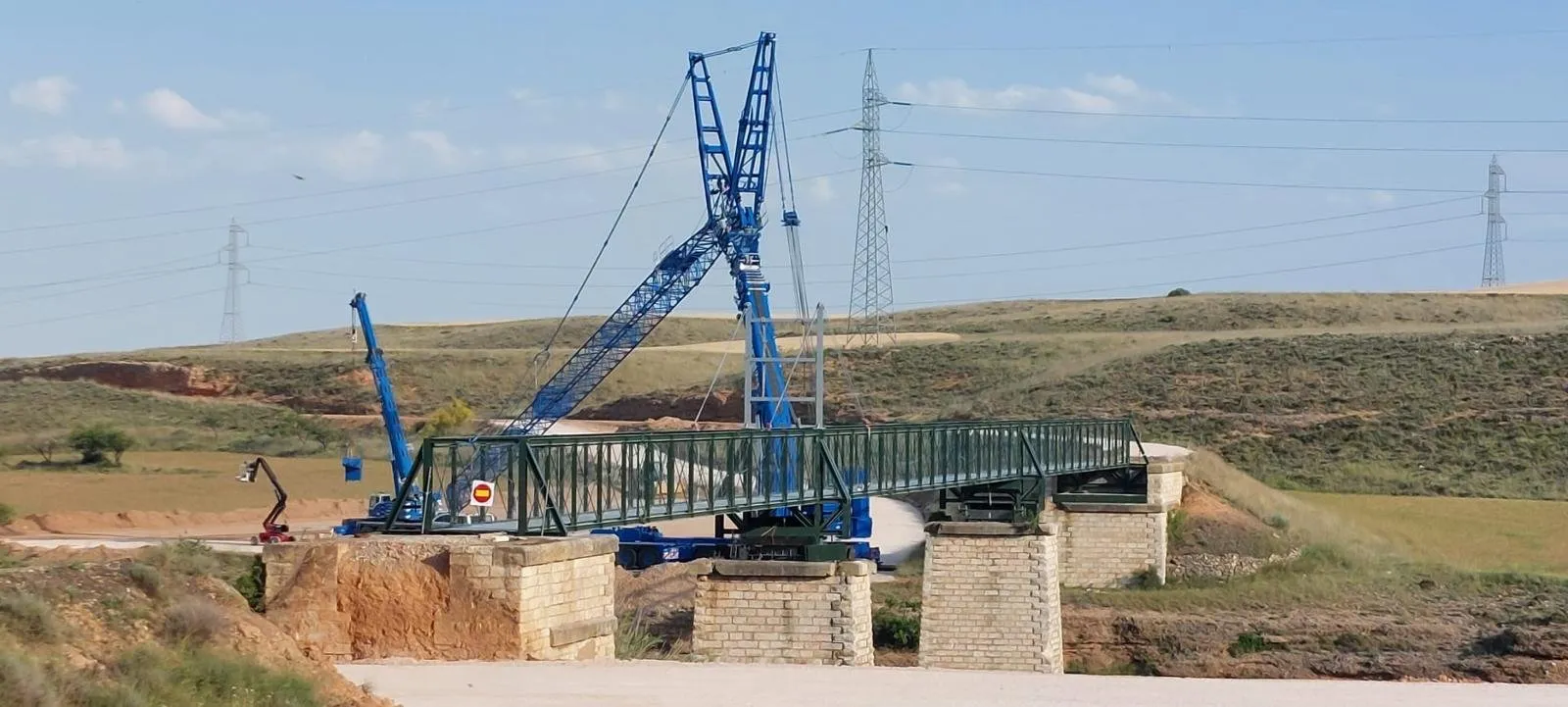 Dos grúas izan la celosía metálica de la pasarela de Belchite